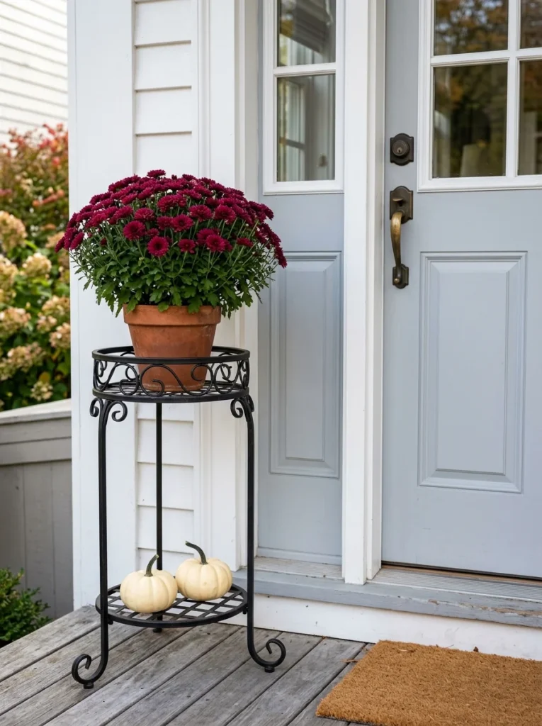 Skinny metal plant stand with mums and pumpkins on a small porch.